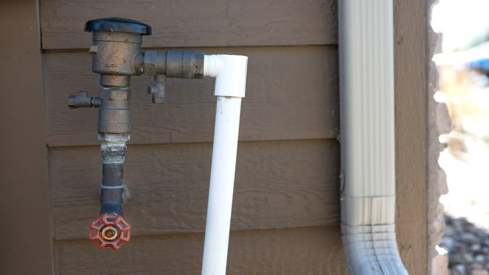 Backflow preventer on a pipe against a house wall beside a downspout