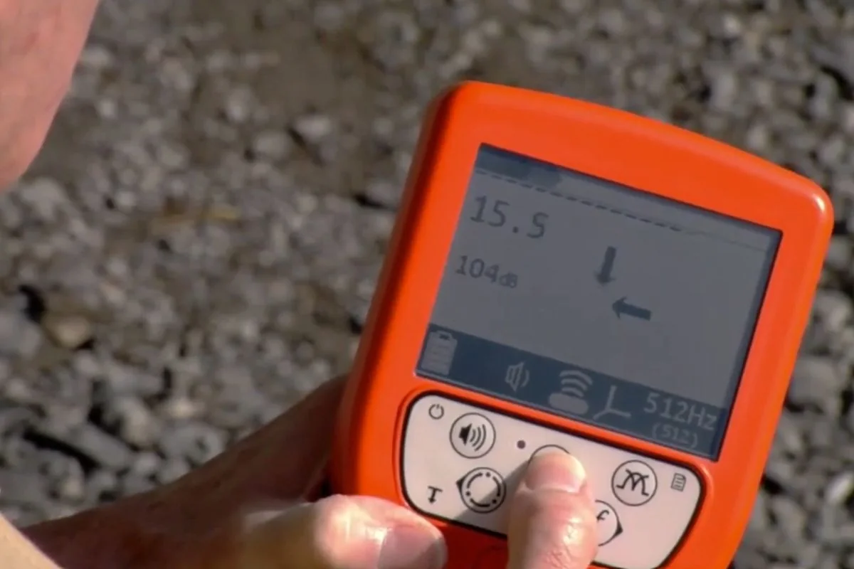 Handheld pipe locator with an orange case showing a depth reading