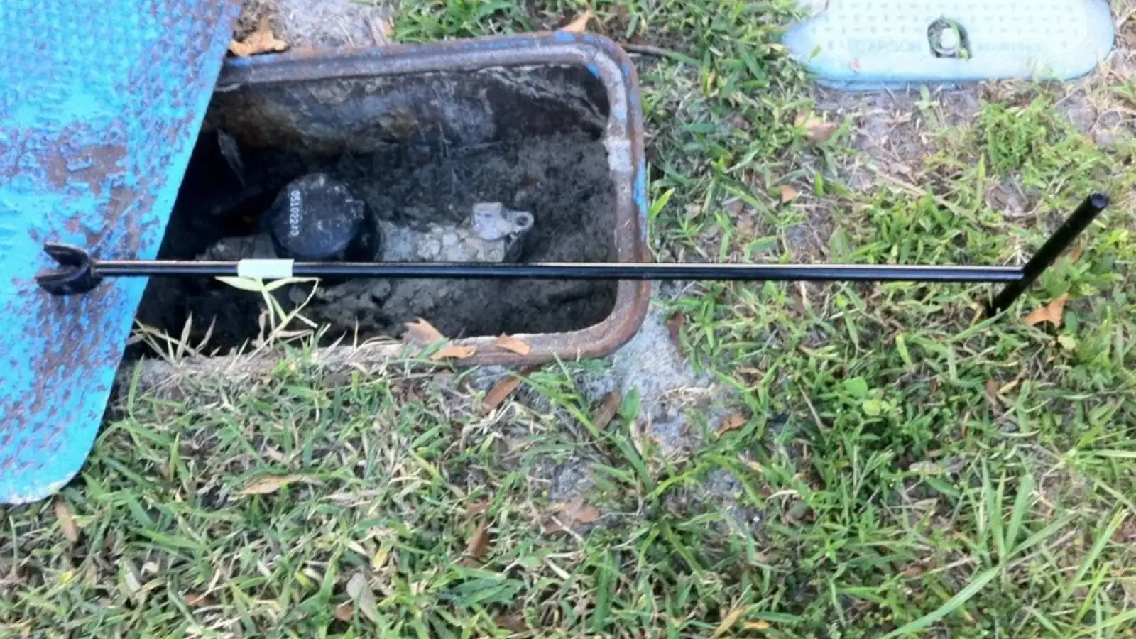 Open meter box in a lawn with a long-handled tool reaching the valve